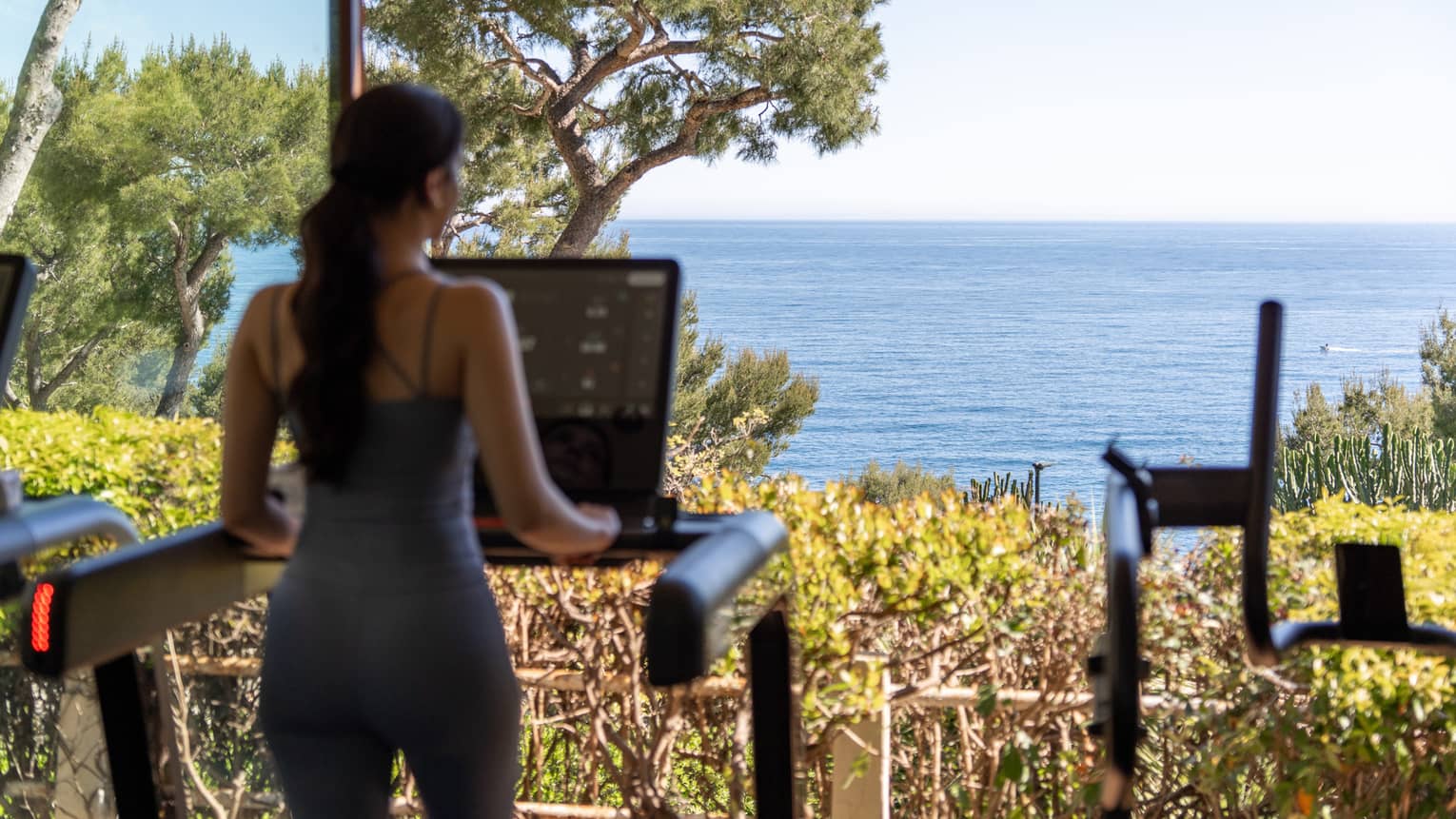 A person stands on a treadmill facing large windows with landscape and ocean views.