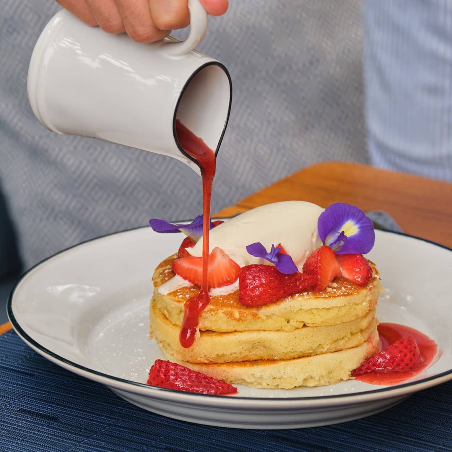 Hand ours red syrup from a white carafe onto a stack of three fluffy pancakes topped with fresh strawberries and cream and garnished with purple flowers