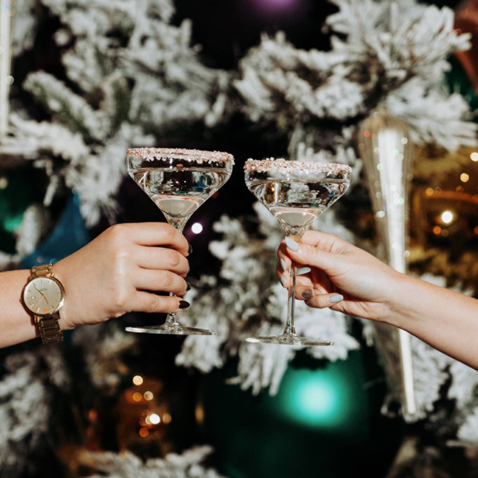 Two hands clink frosted-rimmed martini glasses in front of frosted Christmas tree