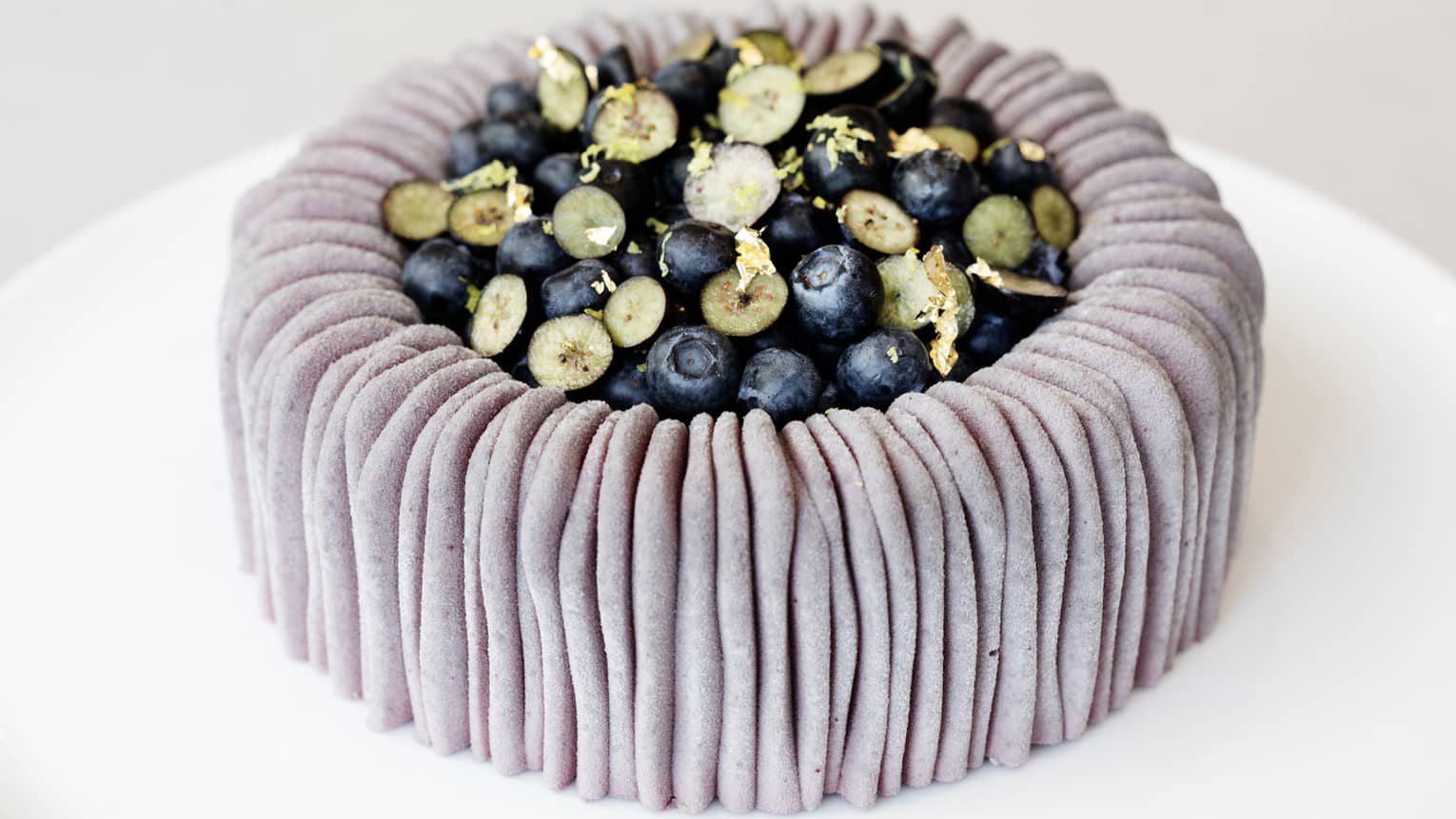 A white plate holding a round cake that has a textured lavender outer edge and is topped with pale green sliced fruit and deep blue berries in the centre