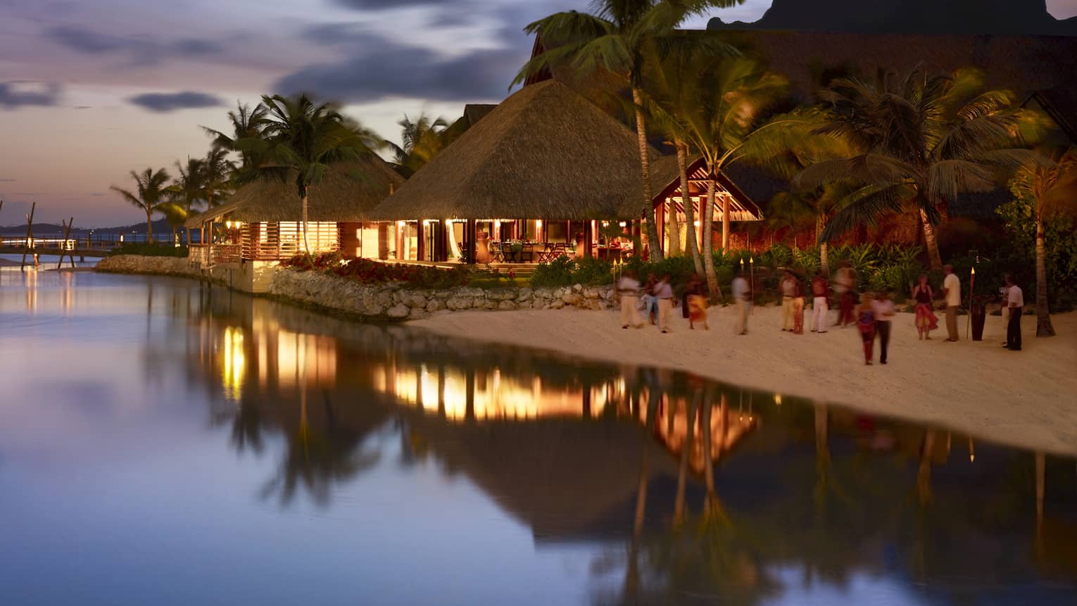 Guests on sand beach by pavilions with lights at night
