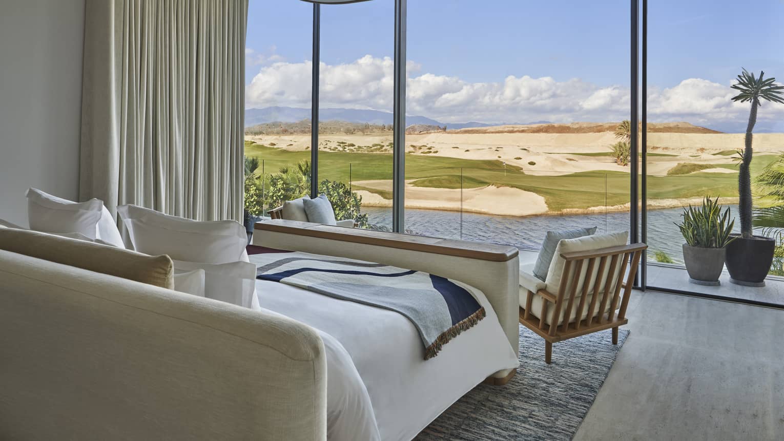 White bed with chair at foot flanked by draped wall and facing tall windows to golf course view