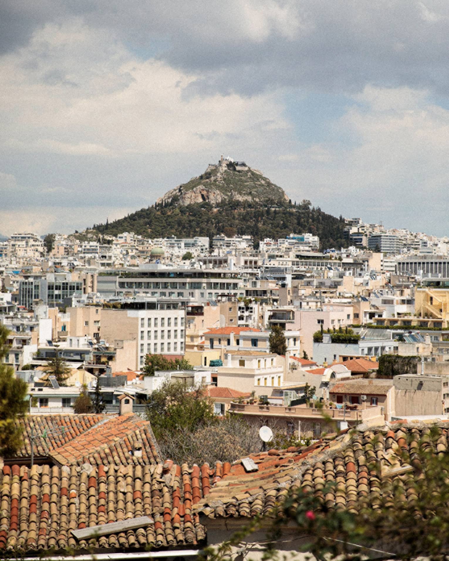 Mount Lycabettus rises from a base of pine trees within the city of Athens, a sprawl of pale modern buildings.