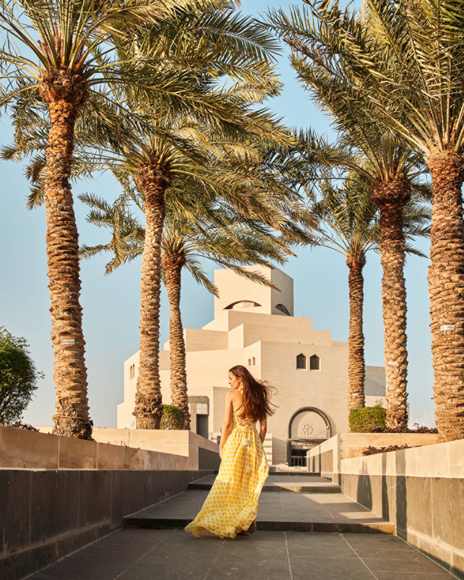 Woman wearing long flowing yellow dress walks along a tiled walkway surrounded by palm trees