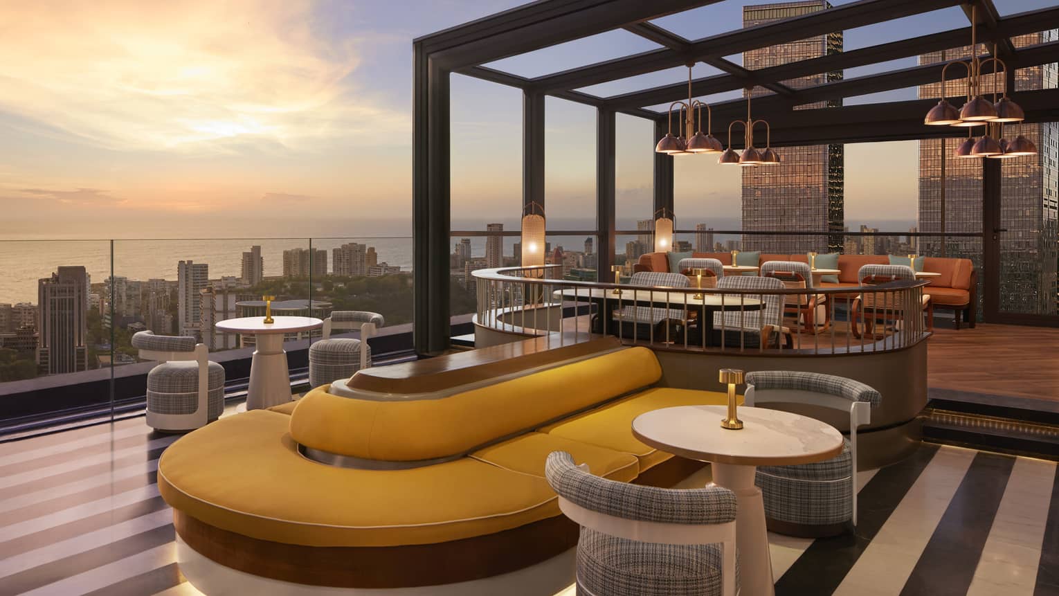 A modern rooftop patio with a mix of booth bank seating and tables for two, panoramic cityscape view and dusky ocean beyond.