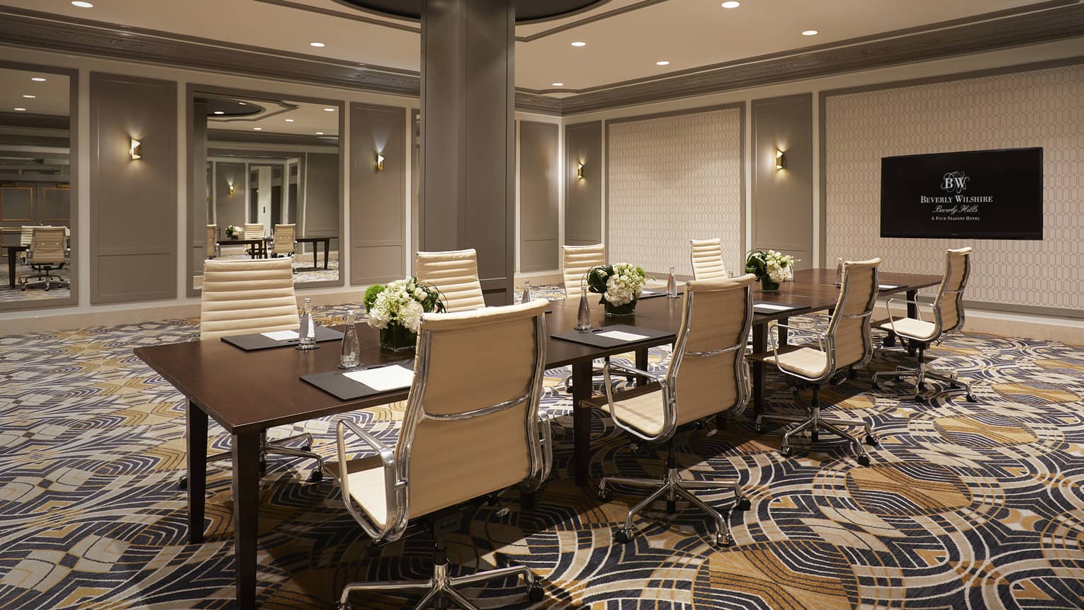 Meeting room with blue, white and yellow patterned carpet and white and grey walls is set up with a rectangular conference table with eight cream chairs and white floral centrepieces