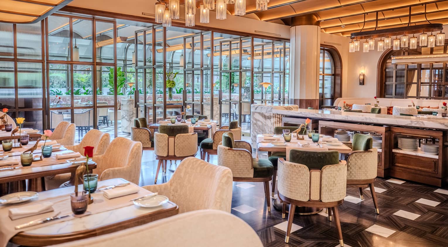 Restaurant dining room featuring tall beamed ceilings, white marble tables, dark flooring and green-and-white velvet chairs all lit by warm overhead lights