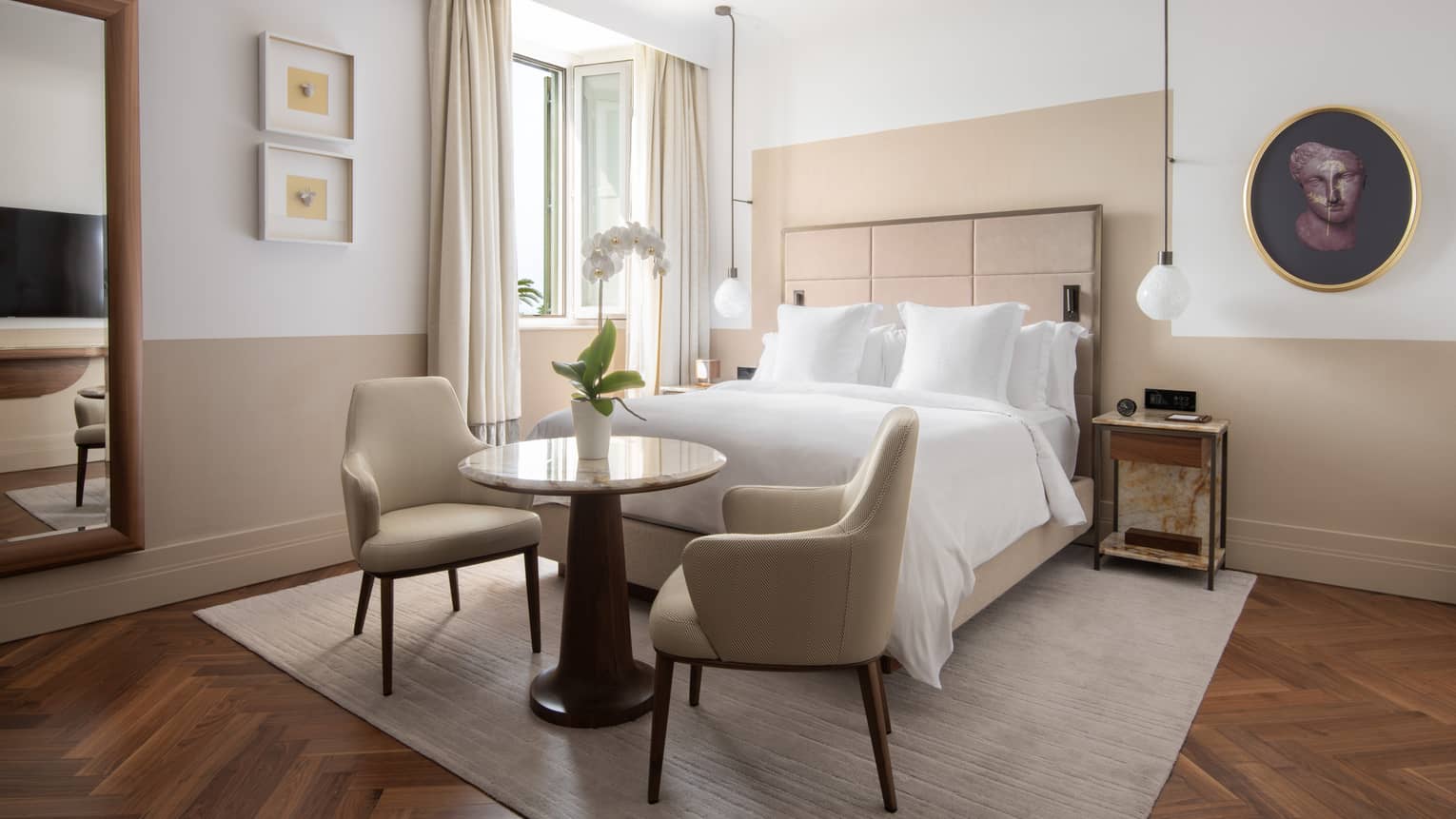 A bedroom with a bed, small table and two chairs, a window, and a mirror.