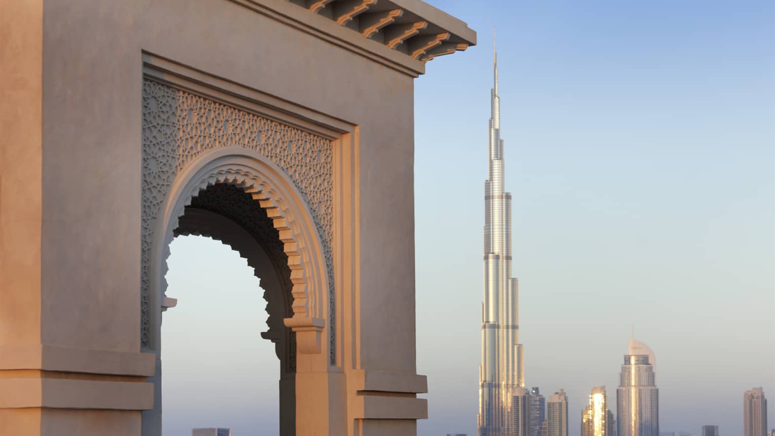 Orange glow of sunset on a decorative stone arch, the iconic Burj Khalifa gleaming in the distance against blue sky.,Orange glow of sunset on a decorative stone arch, the iconic Burj Khalifa gleaming in the distance against blue sky.