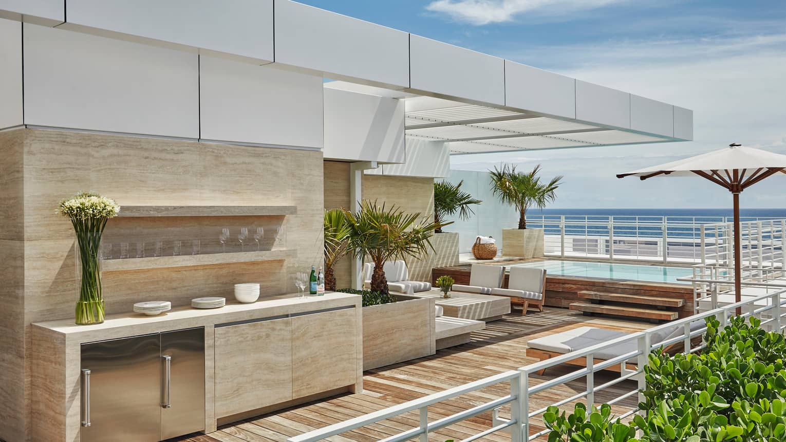 Marble outdoor bar and kitchenette with glasses, flowers, white sofas on modern rooftop patio with ocean views