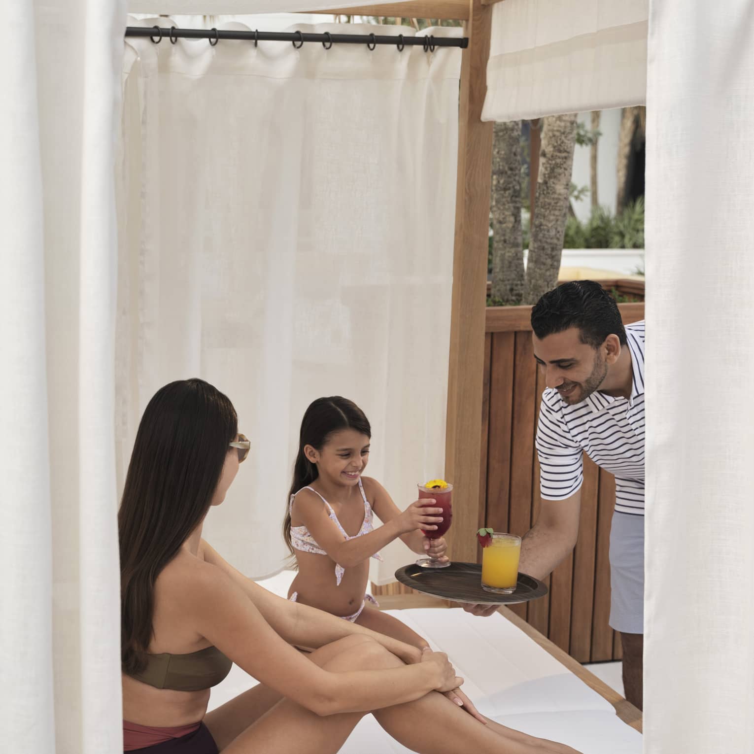 Two guests wearing swimsuits and sitting in a cabana while a person holds a plate with two drinks.