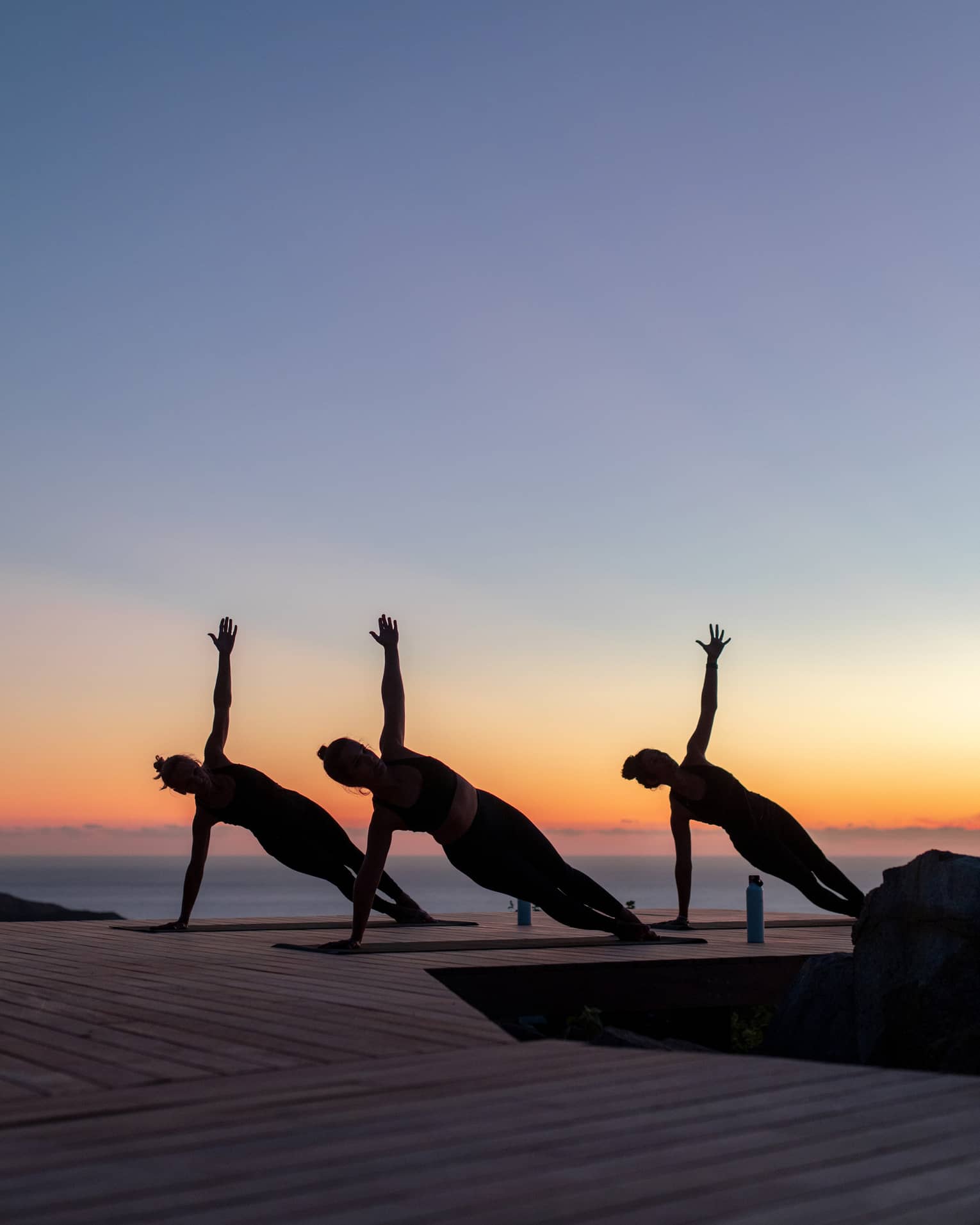 Three guests doing side plank poses on yoga mats on a wooden dock are silhouetted against a clear blue, orange and pink sky.