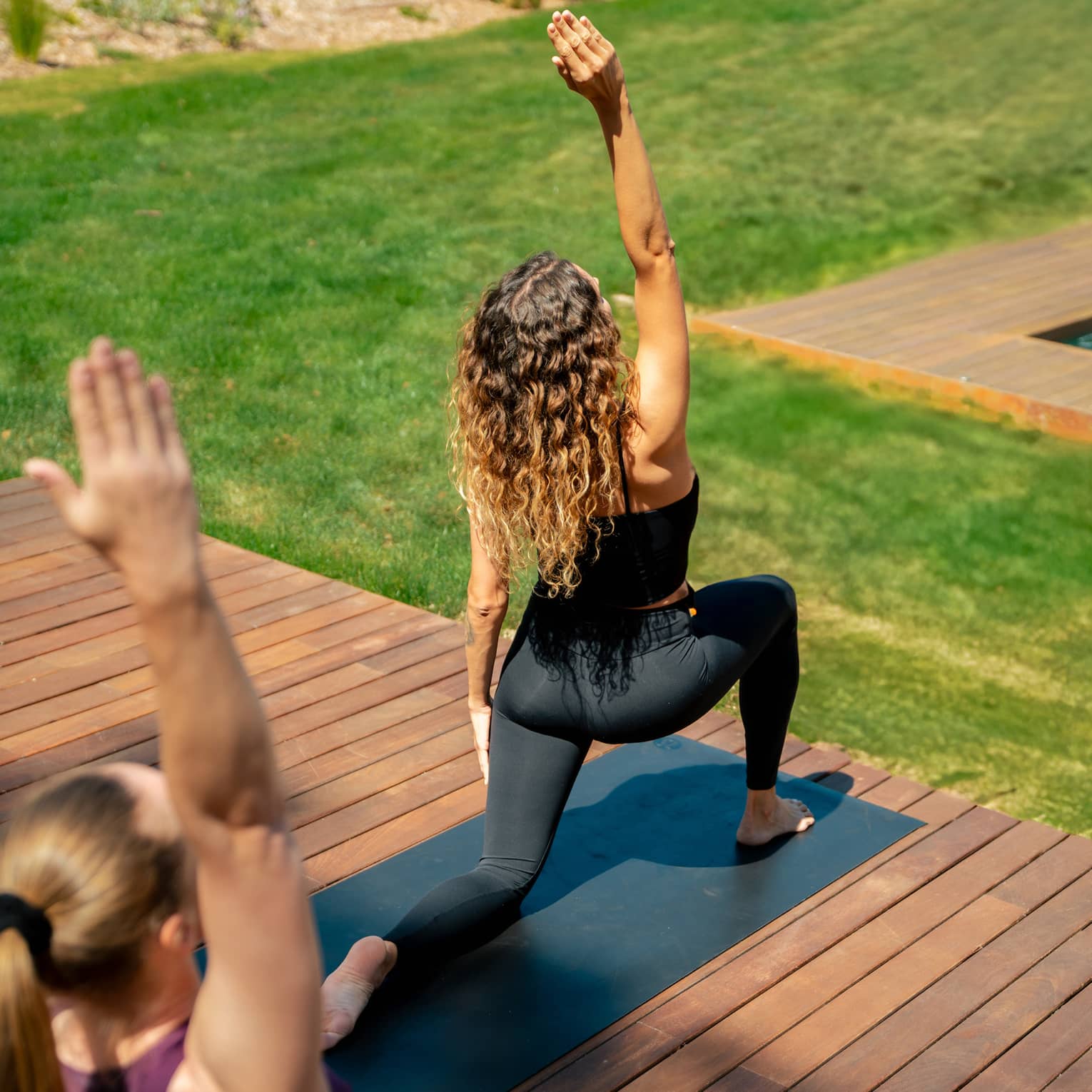 Two people in yoga pose practise on outdoor wooden deck overlooking lush green grass