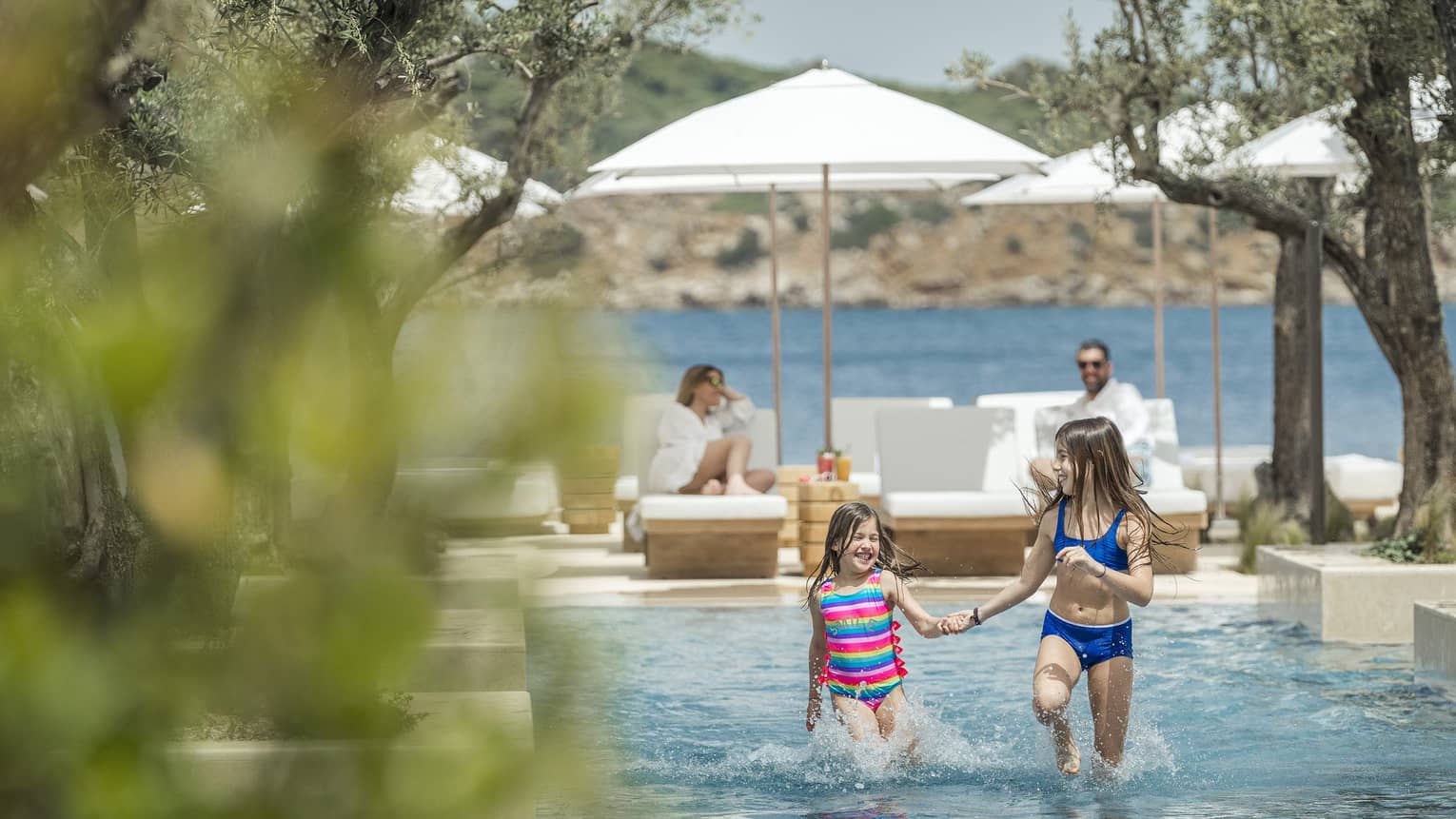 Two smiling children frolick in a shallow outdoor pool while their parents watch from sun loungers. The ocean is behind them.