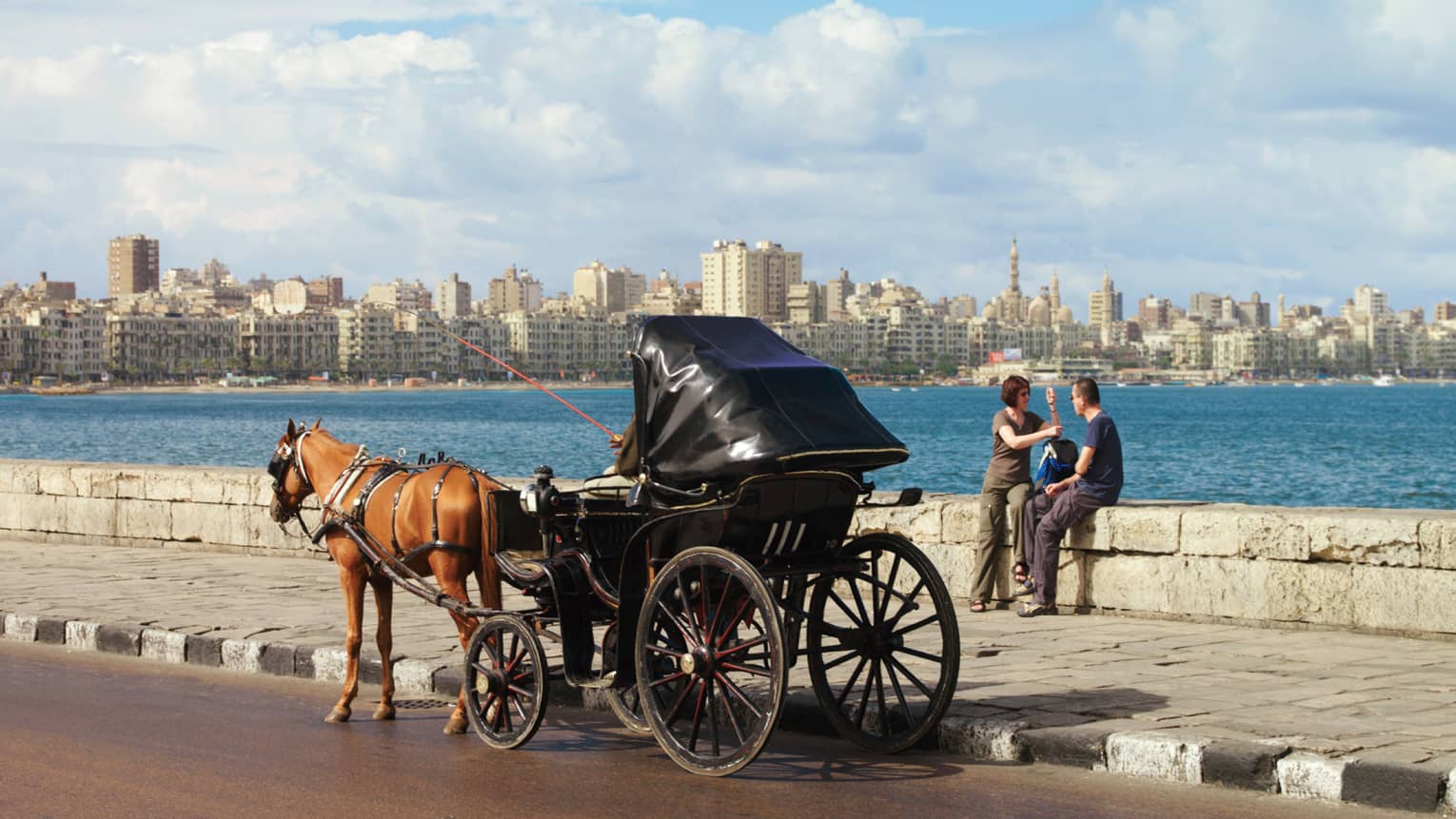 Caleche on the Corniche with couple seated on low rock wall, overlooking water and city skyline