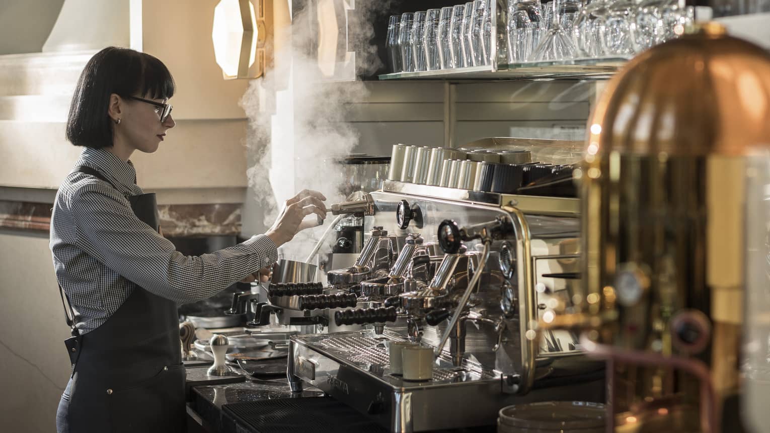 A woman with a bob haircut works at an espresso machine surrounded by crystal glasses. 