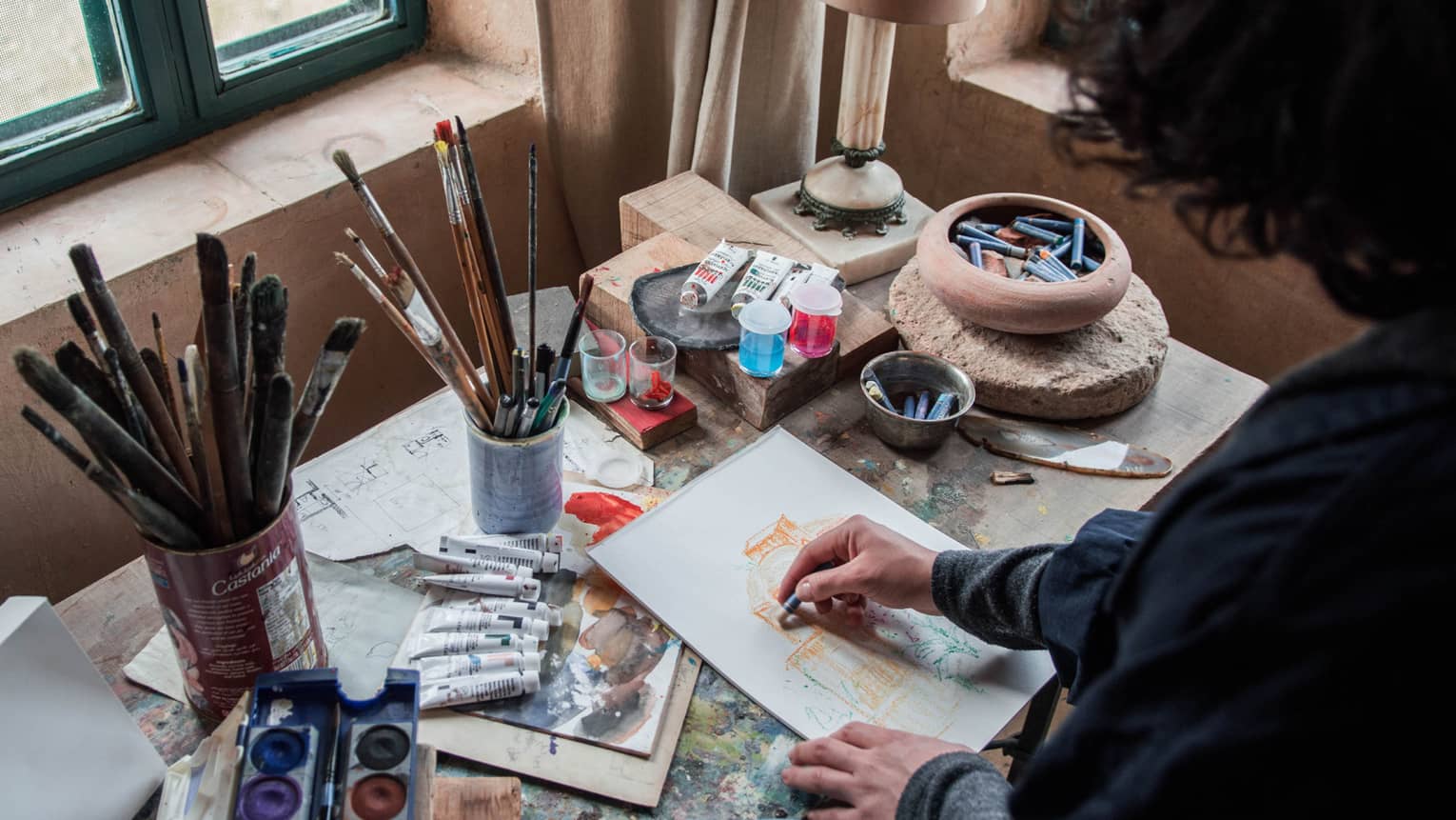 A person with dark curly hair in front of an artists workstation with paintbrushes and acrylic paints.