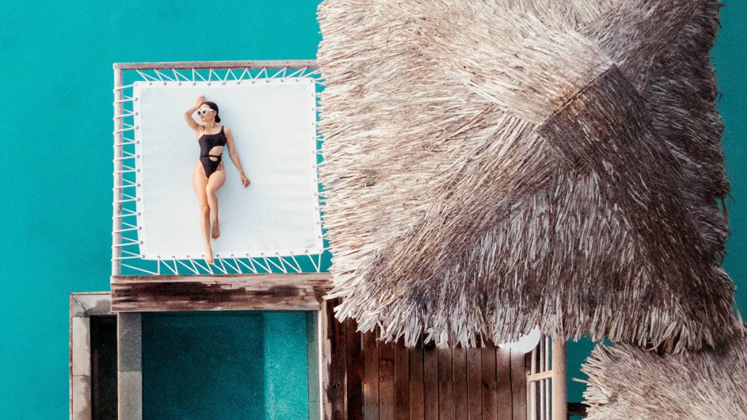 Woman lays on overwater net next to thatched-roof bungalow