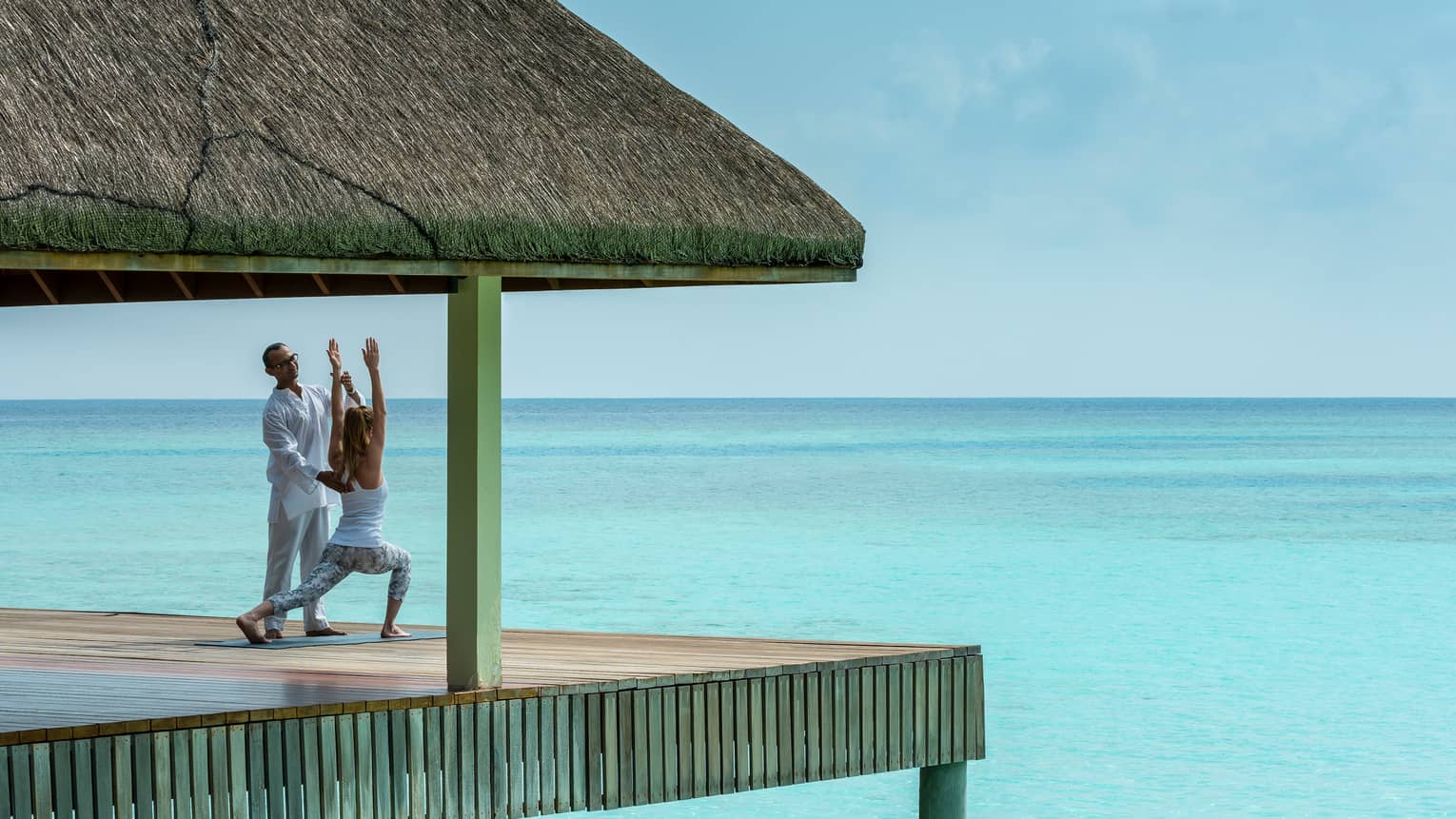 Yogi helps woman into yoga pose on thatched roof pavillion over blue ocean