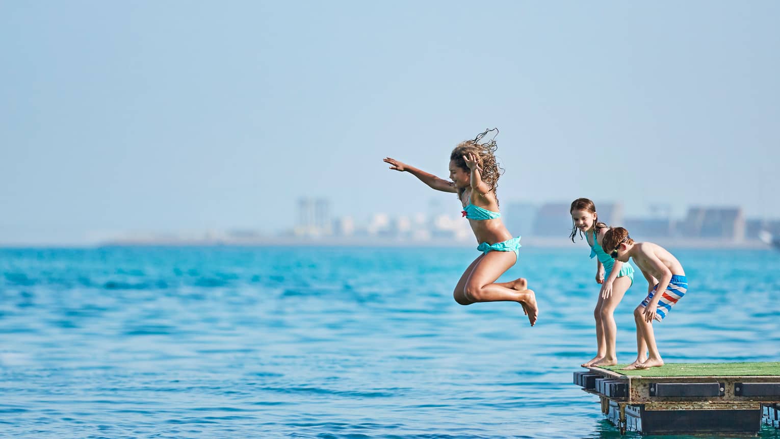 Little girl jumps into sea, two children stand behind her on wood platform