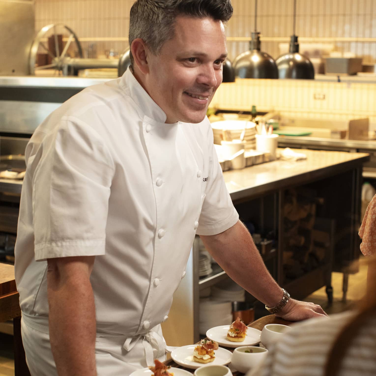 Chef standing beside a dining table in the kitchen talking to guests