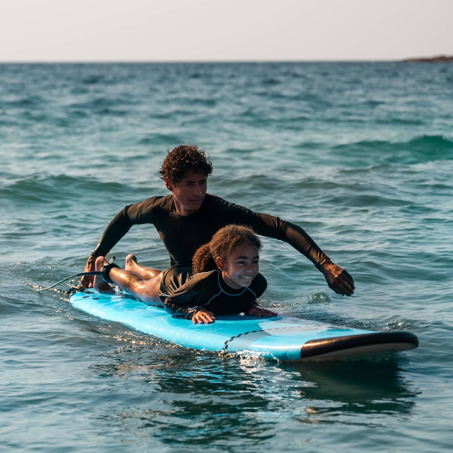 Male suf instructor wearing a black long-sleeve rashguard helps a young surfer paddle in the ocean
