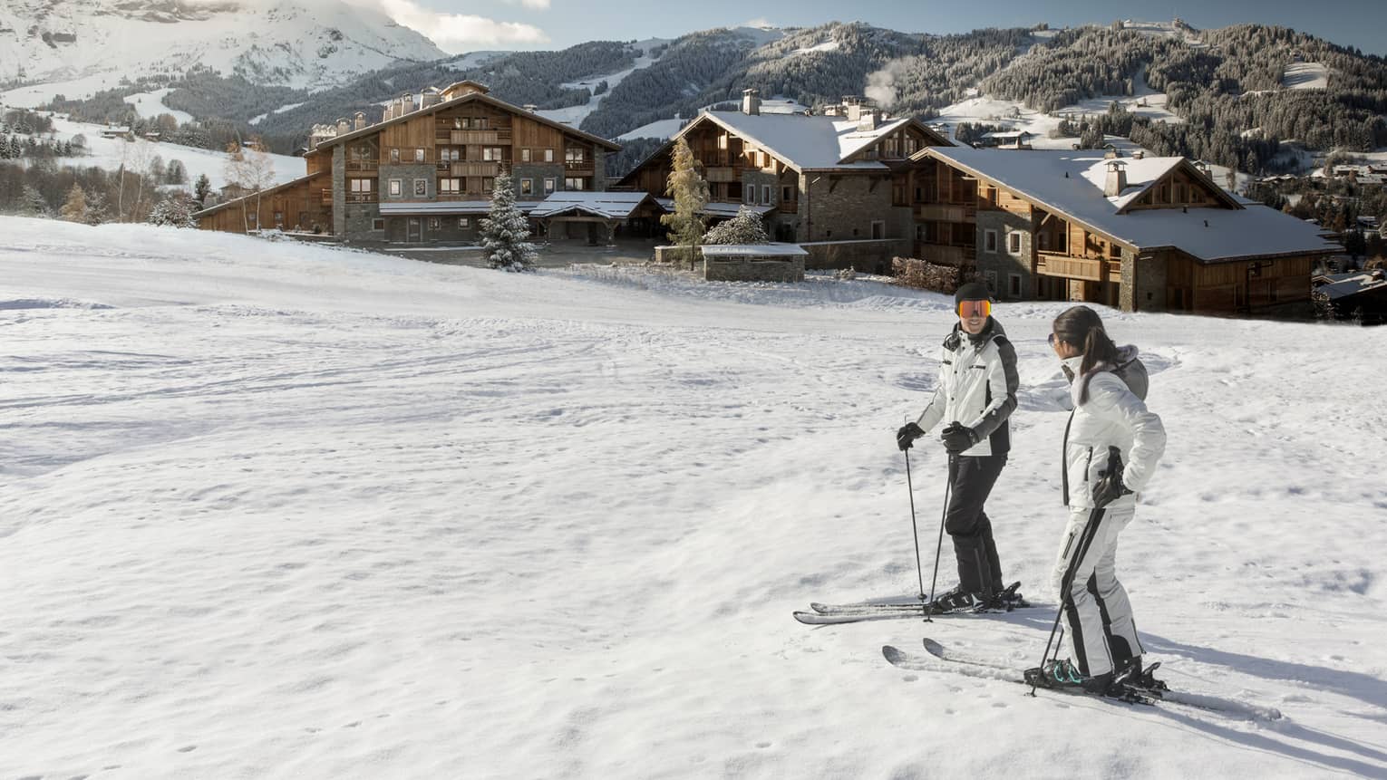 Man, woman on skis at top of snowy hill, Four Seasons Hotel Megeve in background