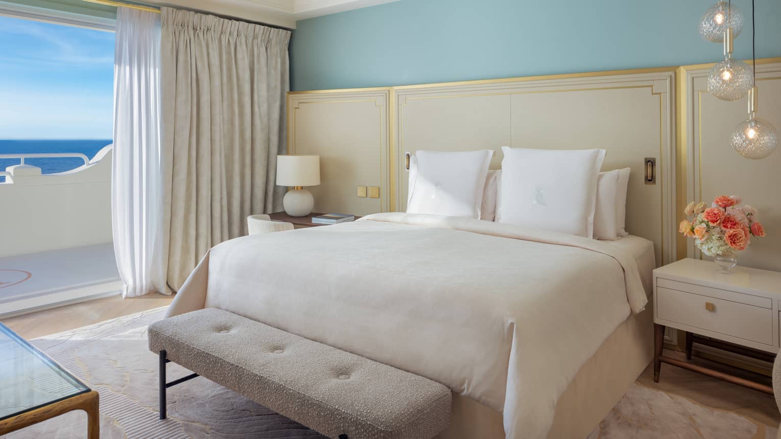 Elegant luxury hotel suite bedroom with king bed, ivory bedding and headboard with gold accents and turquoise wall, with walk-out sea-view balcony