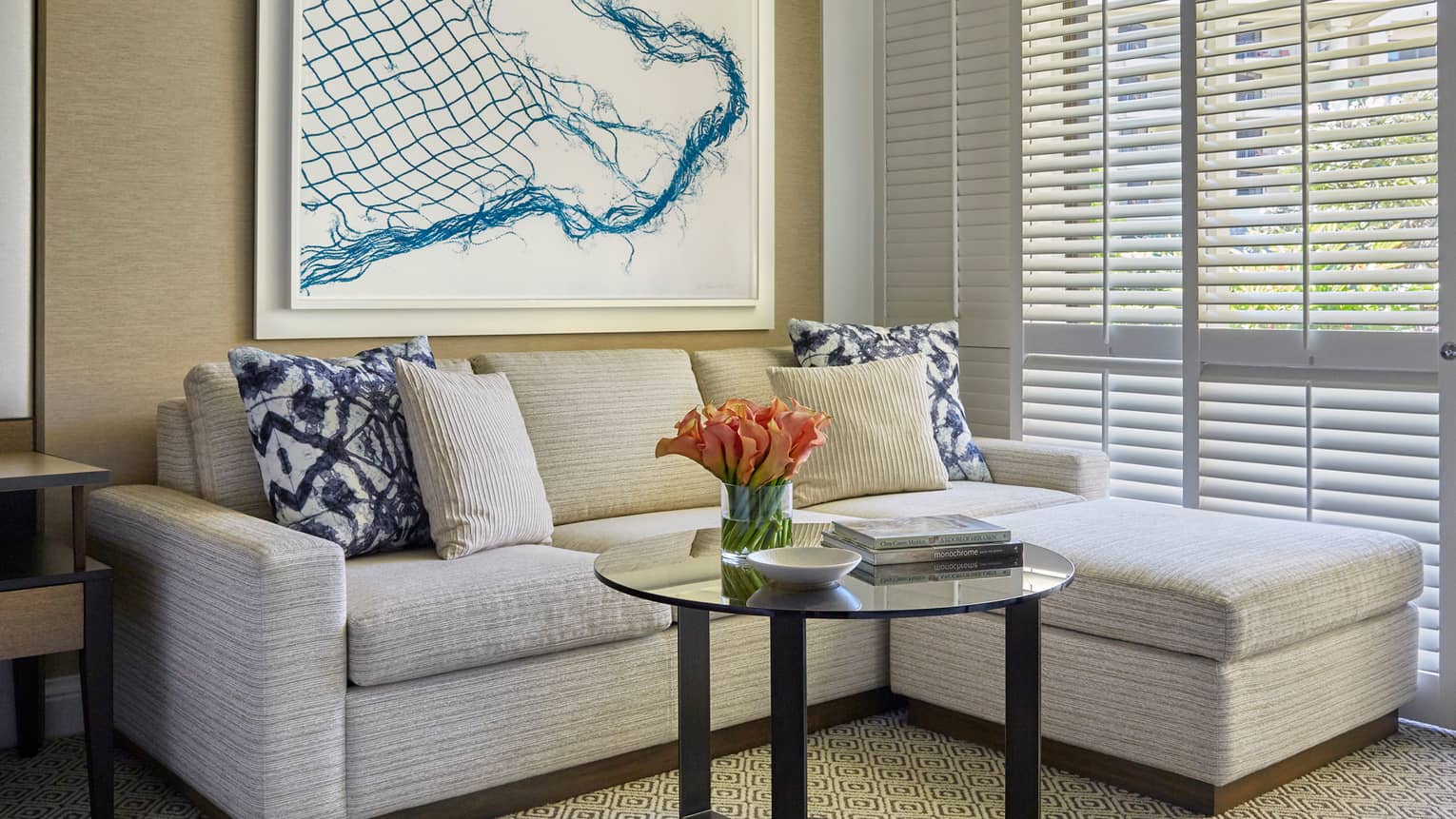 Modern sitting area with light sectional sofa, patterned pillows, glass coffee table and abstract art near window blinds