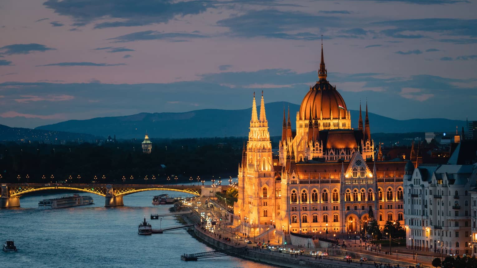Illuminated bridge spanning a river at dusk, with a grand, lit-up building and boats along the waterfront.