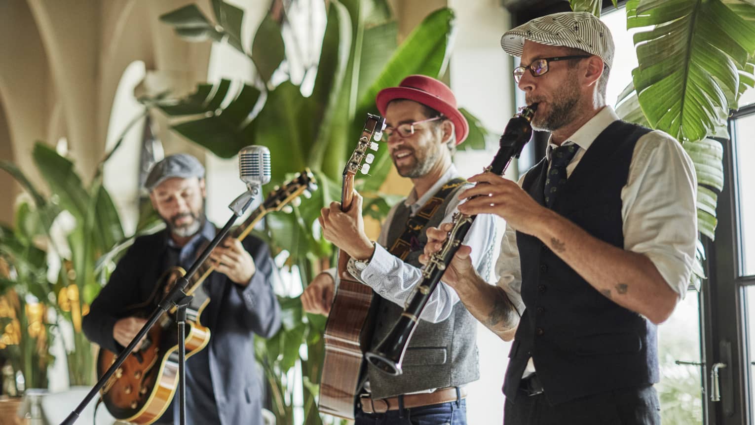Two guitarists and an oboe player stand in front of a wall of tree-lined windows