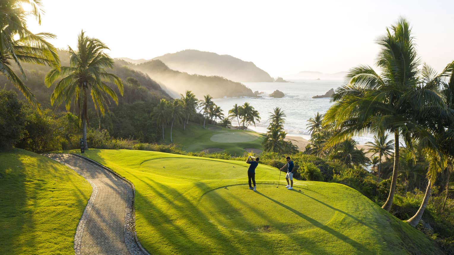 Rear view of two golfers on an elevated tee, one swinging toward the green down by the beach, misty hills and ocean beyond.