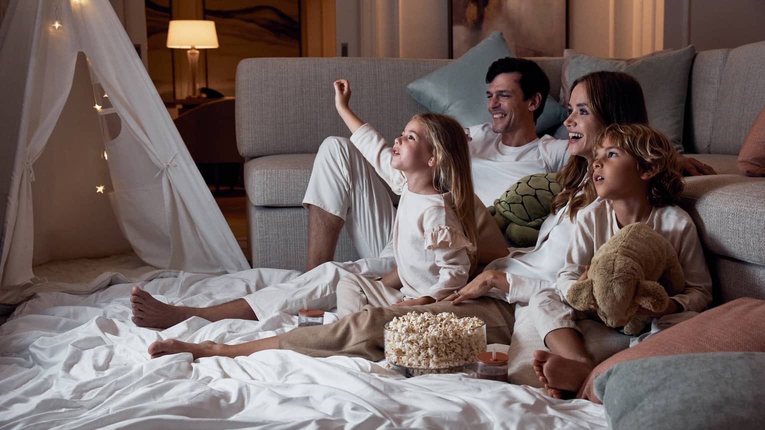 A family relaxing on blankets beside a sofa; the children hold stuffed toys and there's a bowl of popcorn between them.