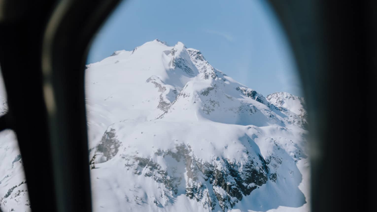 An aerial view through an airplane window of a mountain covered in snow with sporadic clumps of trees around the base.