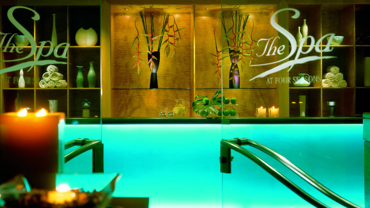 Glass doors at spa lobby, reception desk with with glowing candles, rolled up white towels on shelf 