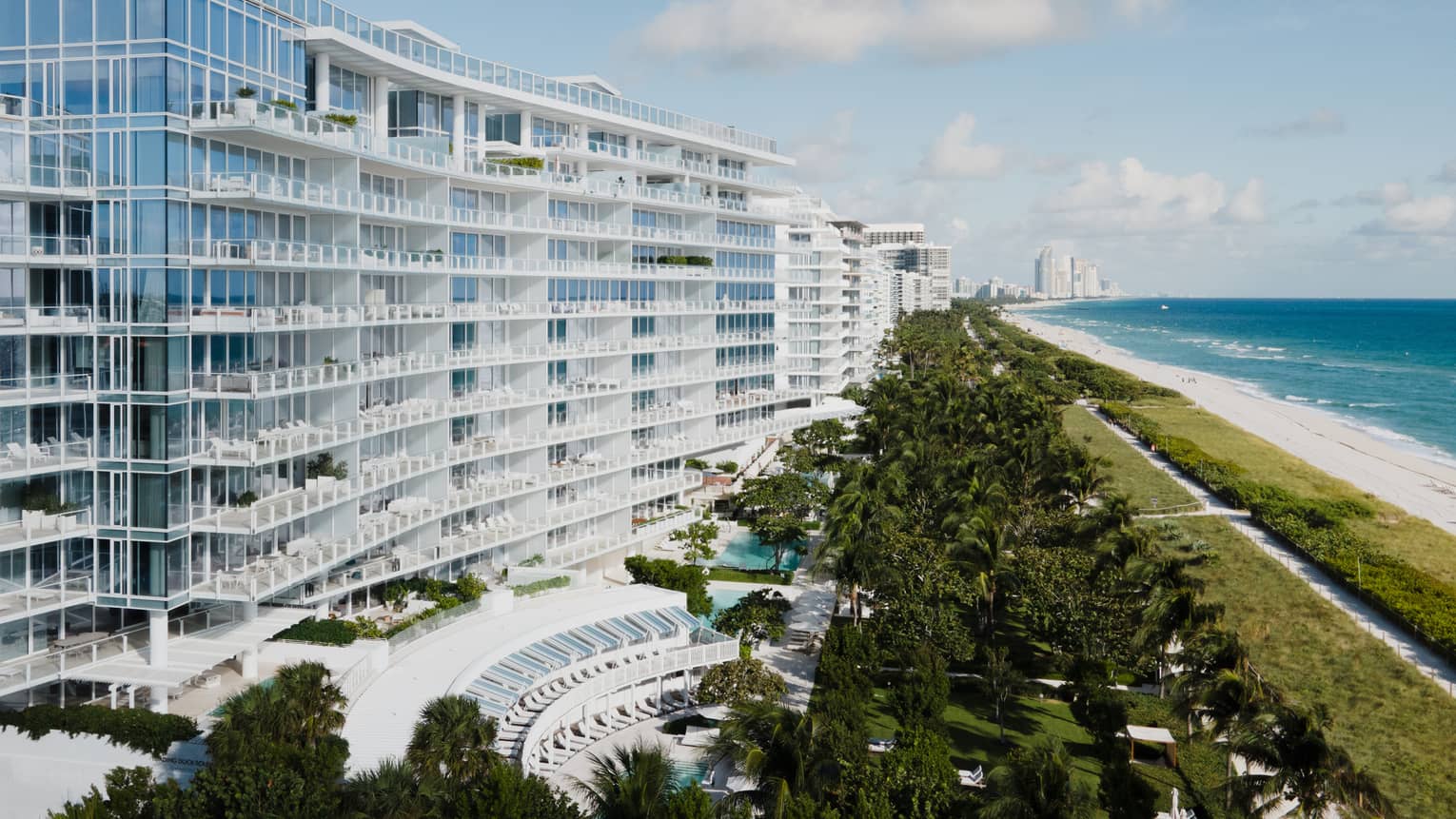 Luxury beachfront hotel with modern glass architecture, lush greenery, a pool area and a view of the ocean and sandy beach under a blue sky