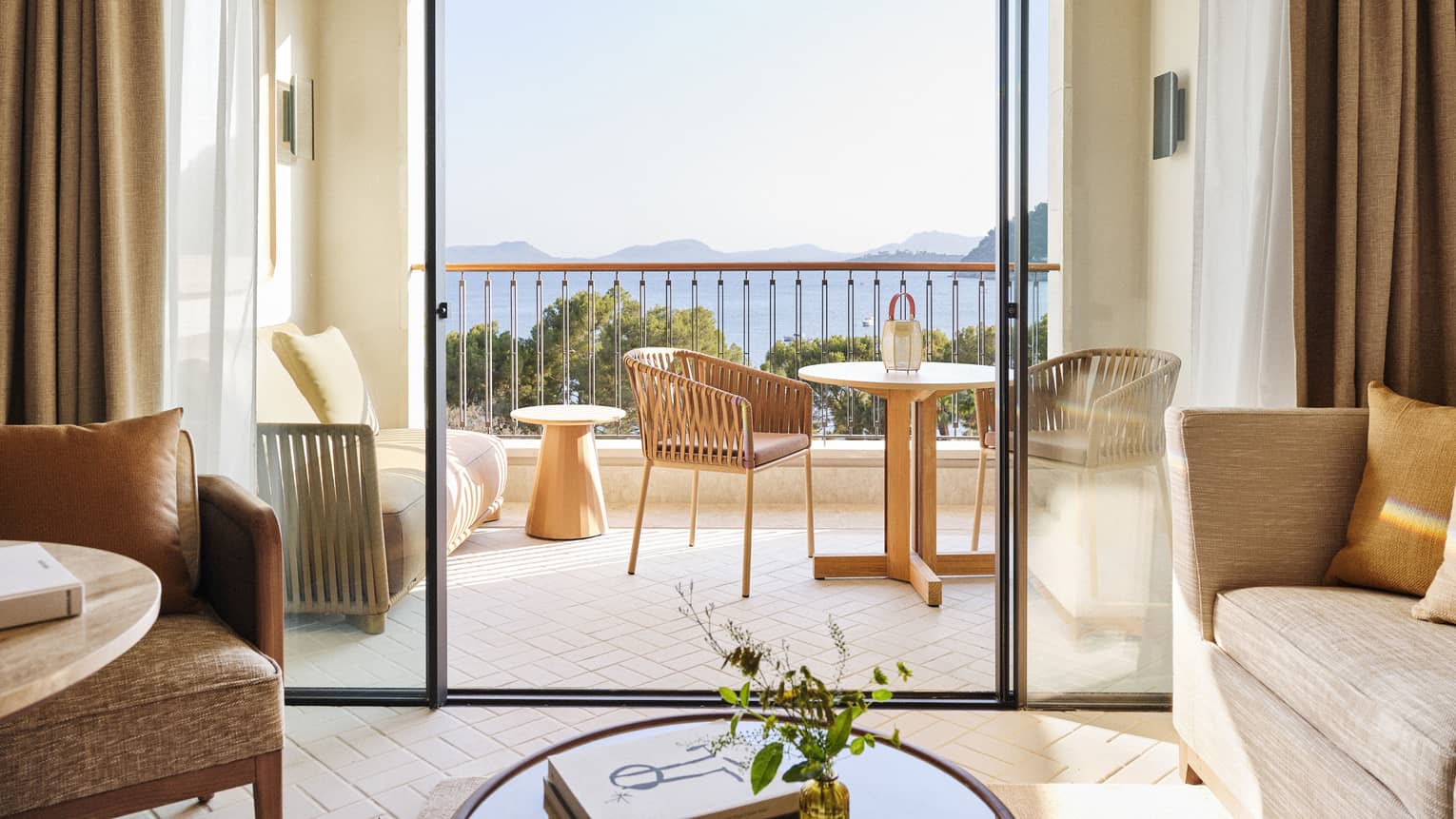 Guest room view of a neutral bistro table and love seat on a sunny balcony overlooking treetops and the ocean beyond.