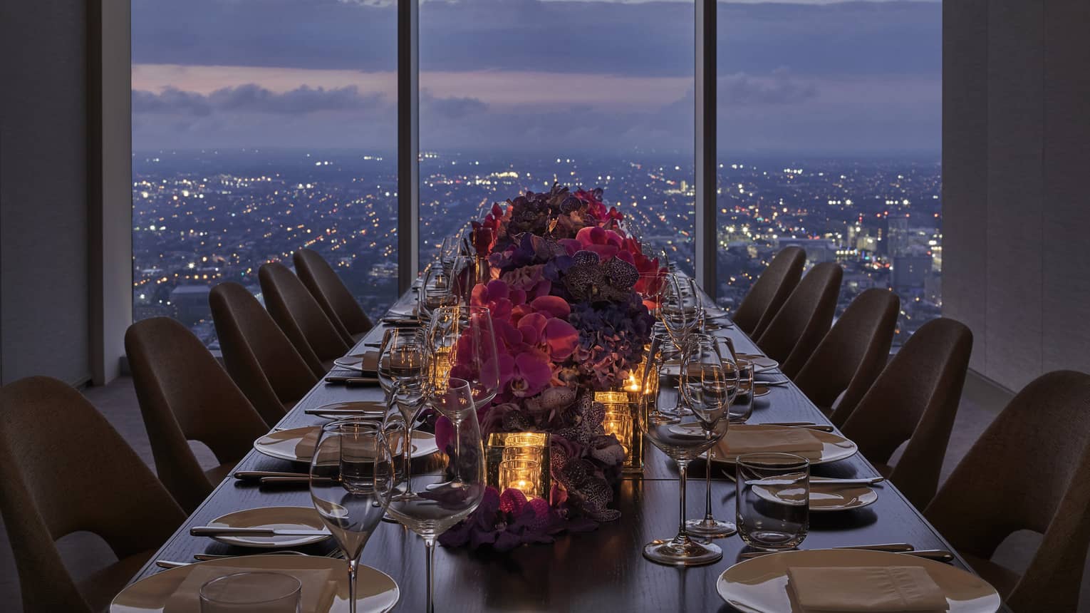 A long dining table with red flowers in the center, a sun set can be seen above a city through a large window.