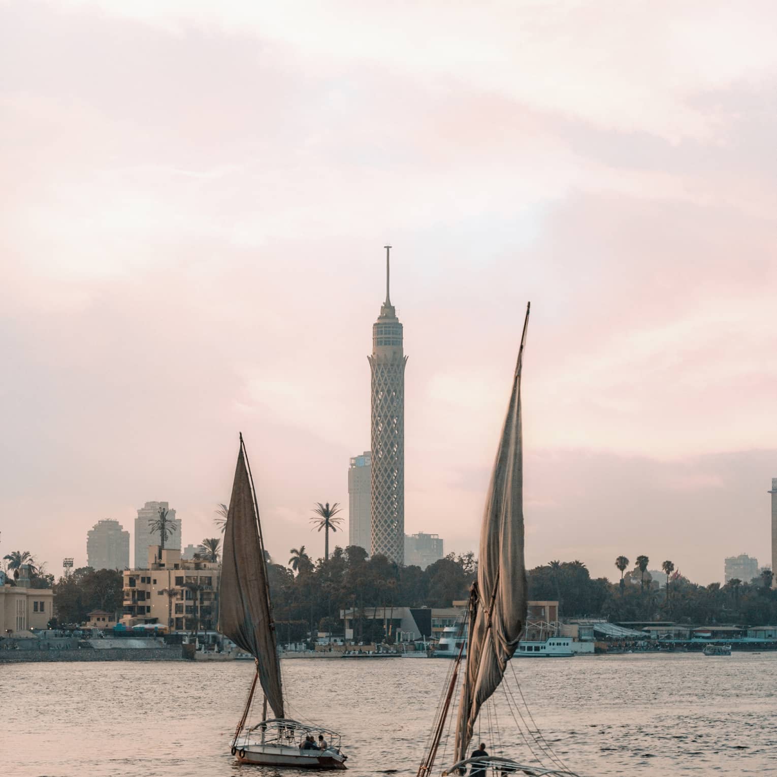 Two felucca boats drift on the water in the evening light, a busy shoreline and Cairo Tower in the distance. 