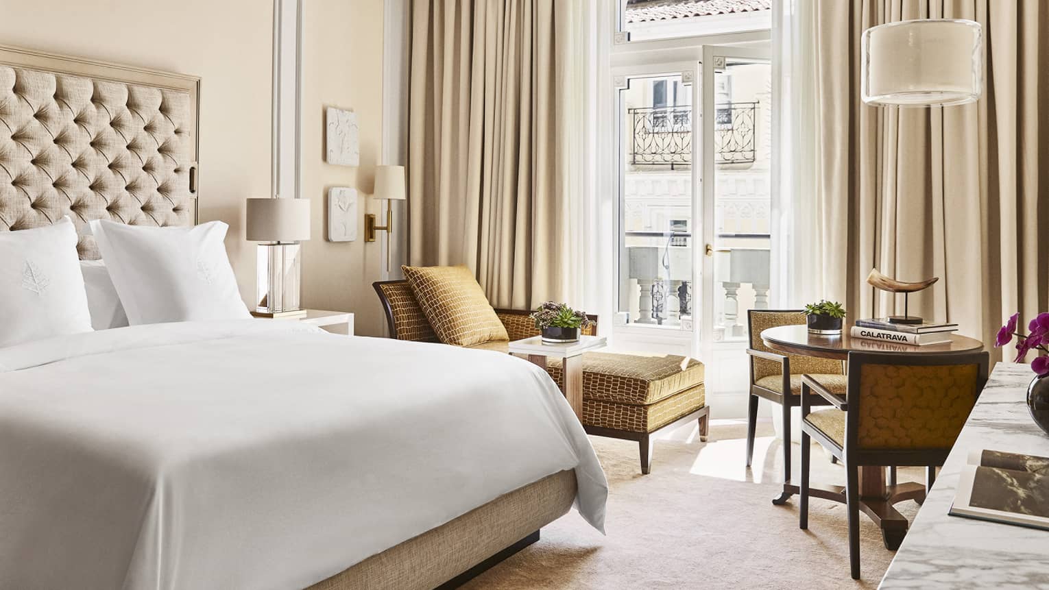 Luxury hotel bedroom with king bed, tufted headboard, table with books and horn sculpture, tan chairs and chaise lounge and doors opening to a Juliet balcony