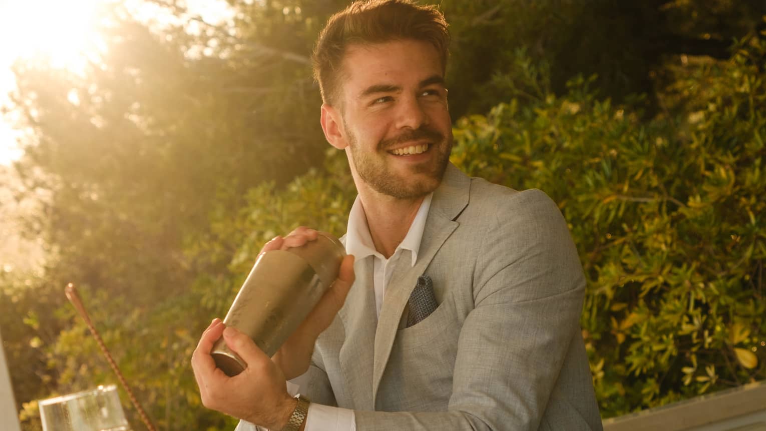 A man shakes a cocktail shaker behind a bar as the sun shines through the trees