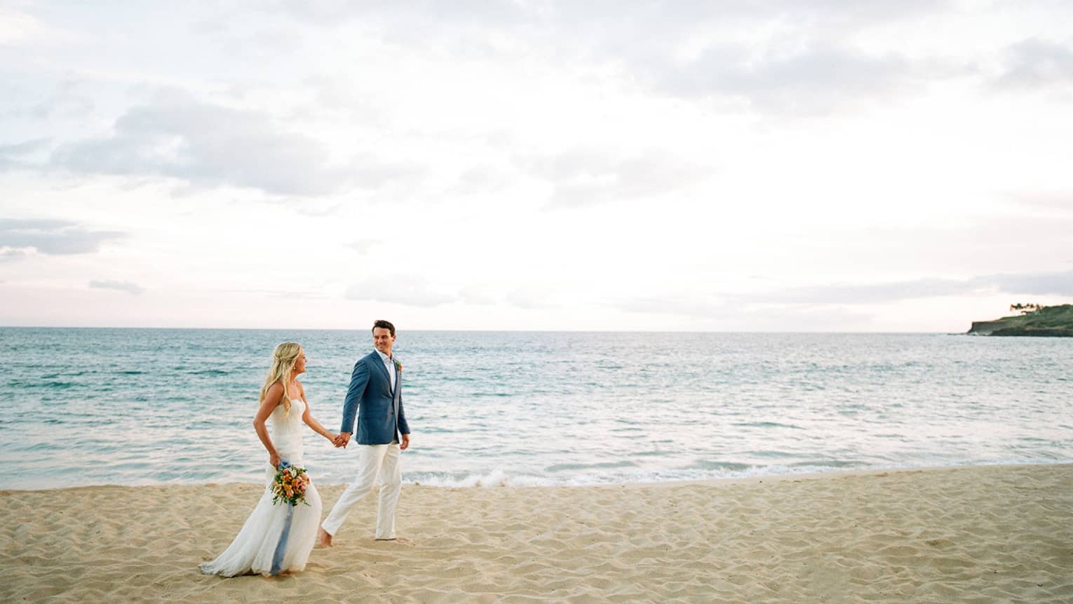 A groom is holding a brides hand as they walk along a beach.