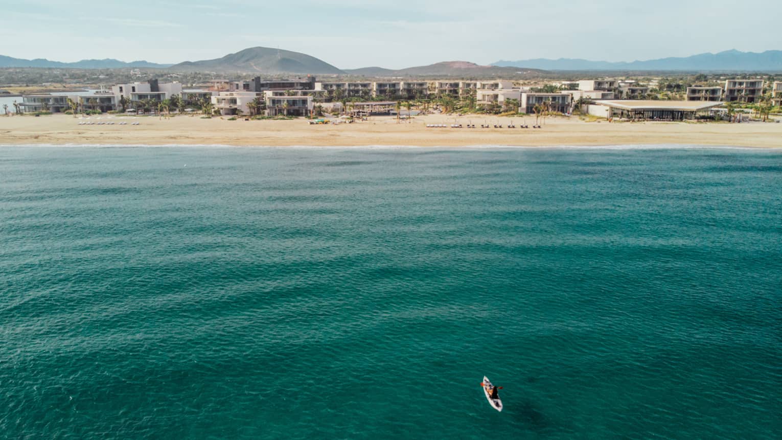 A person on a surfboard out on green-blue water, buildings can be seen in the distance on the shore.