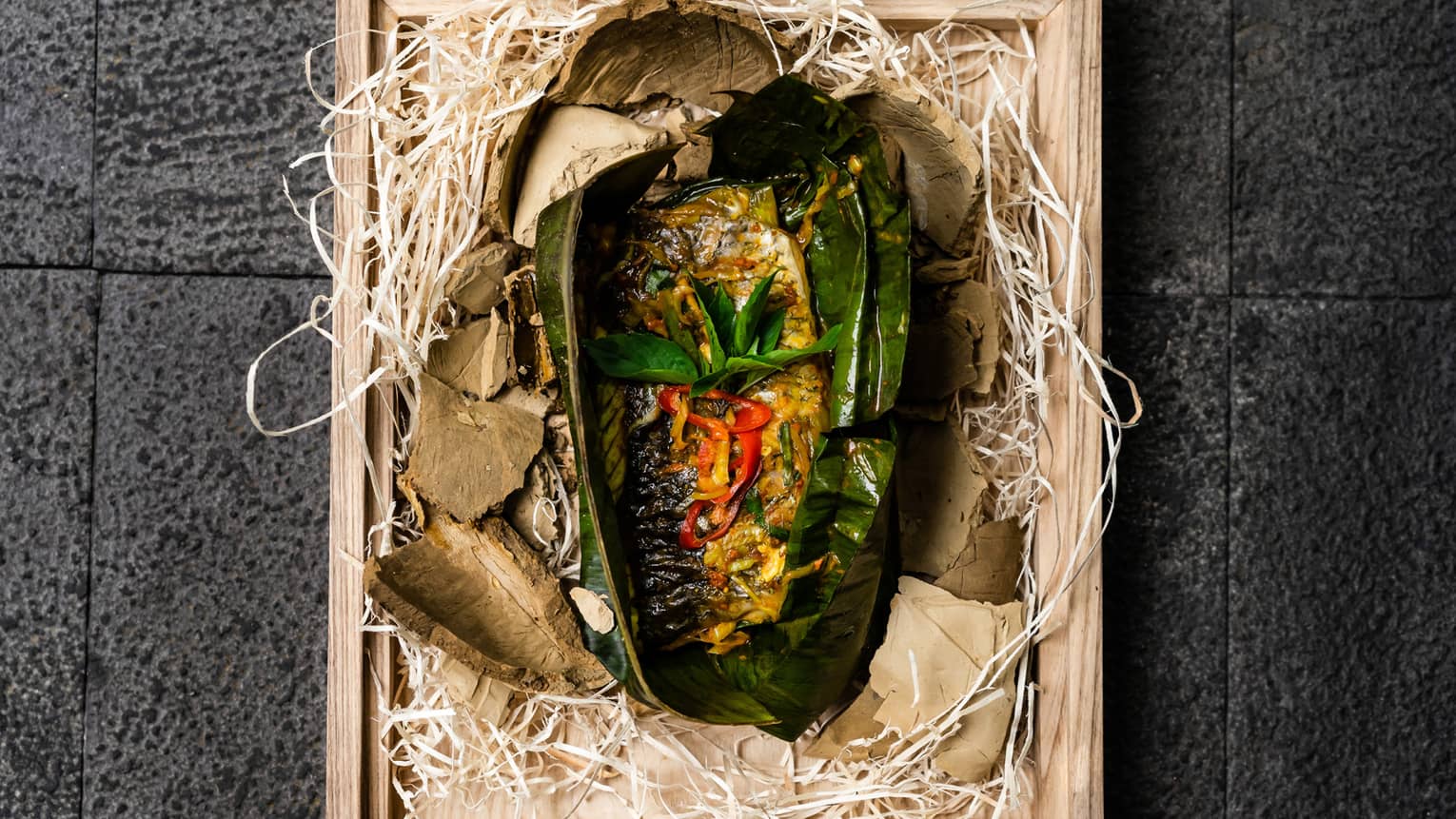 Aerial view of clay-baked sea bass fillet wrapped in banana leaves on wooden tray