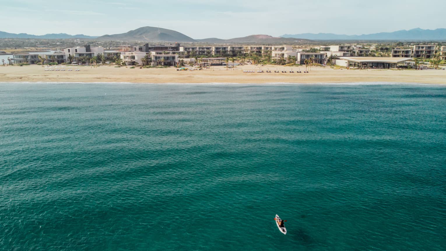 A person on a surfboard out on green-blue water, buildings can be seen in the distance on the shore.