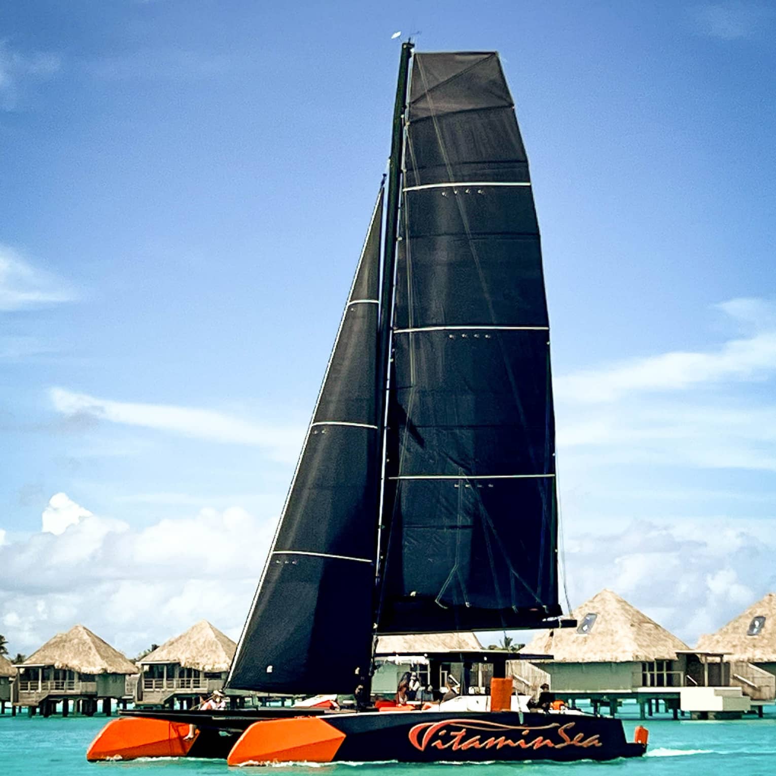 A catamaran named "Vitamin Sea" sails through turquoise waters past a row of thatched-roof overwater bungalows.