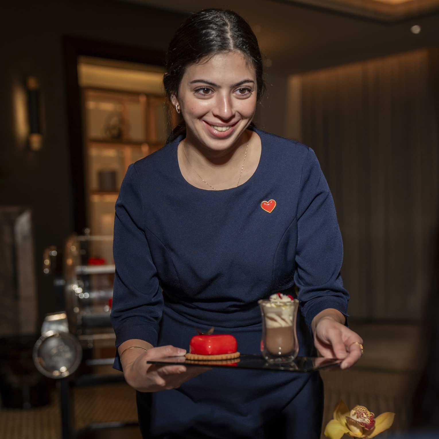Hotel team member wears a navy blue uniform with a red heart-shapped pin as they serve a guest a red heart-shaped cake and hot chocolate