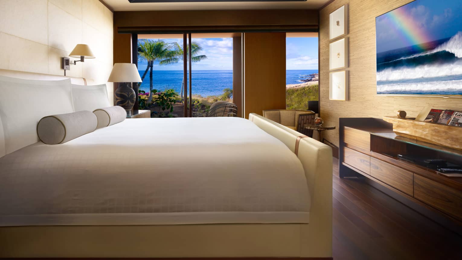 Four Seasons Oceanfront Studio Suite bed with white padded frame, headboard, dresser under TV displaying waves, sunset