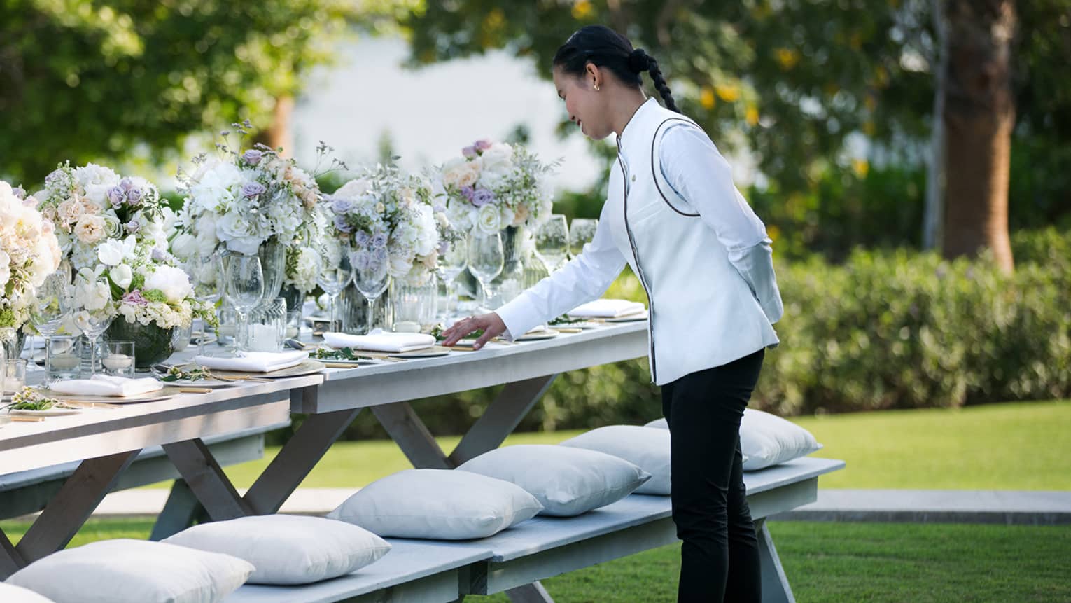 A four seasons staff does the final touches on an outdoor, long banquet table decorated with large white floral arrangements 