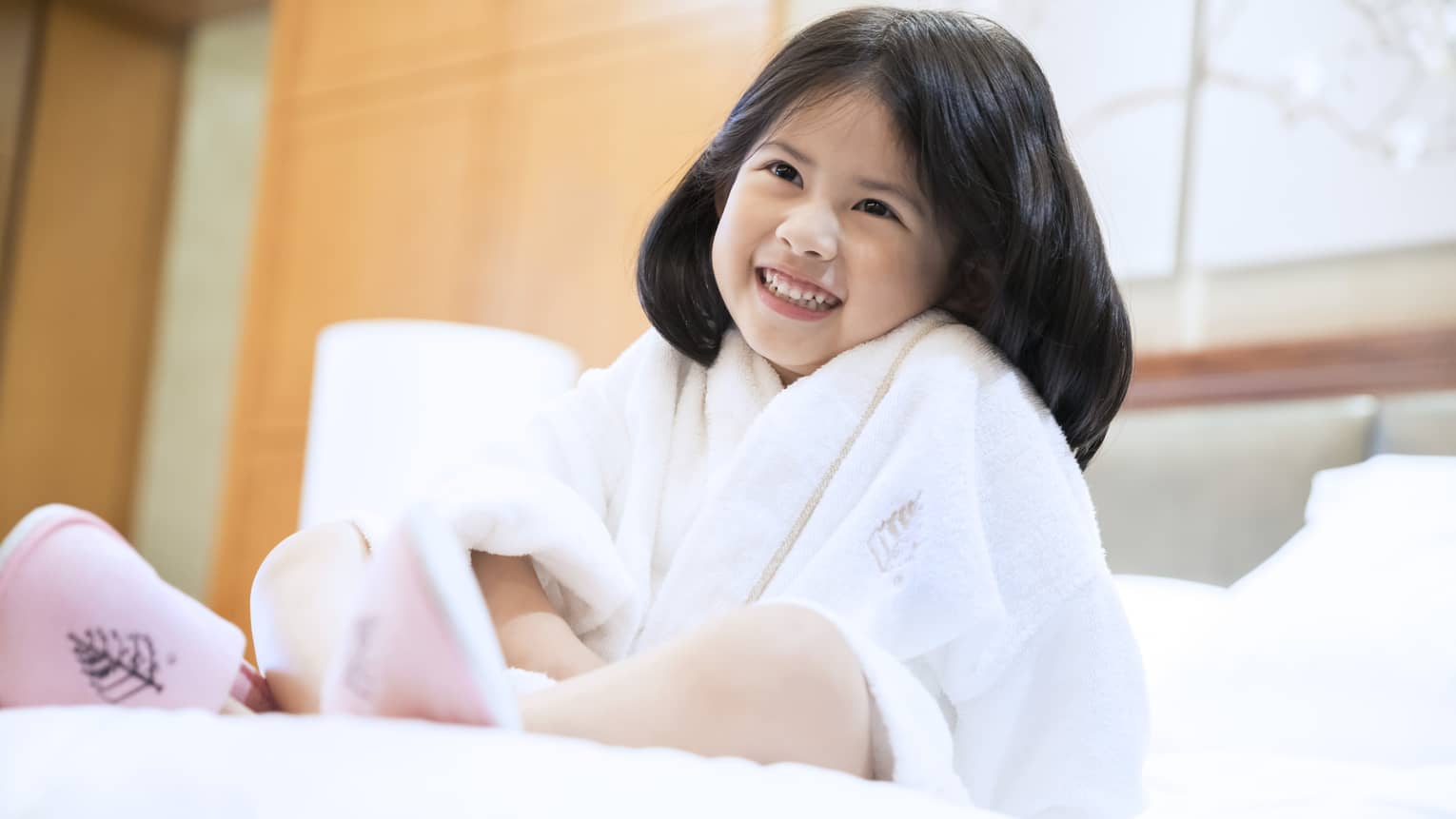 Smiling little girl wearing white bath robe and pink slippers sits on bed